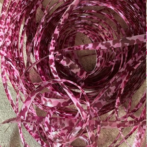May include: A close-up of a coil of pink camouflage ribbon. The ribbon is a vibrant pink with a darker pink and brown camouflage pattern. The ribbon is curled and twisted, creating a visually interesting texture.