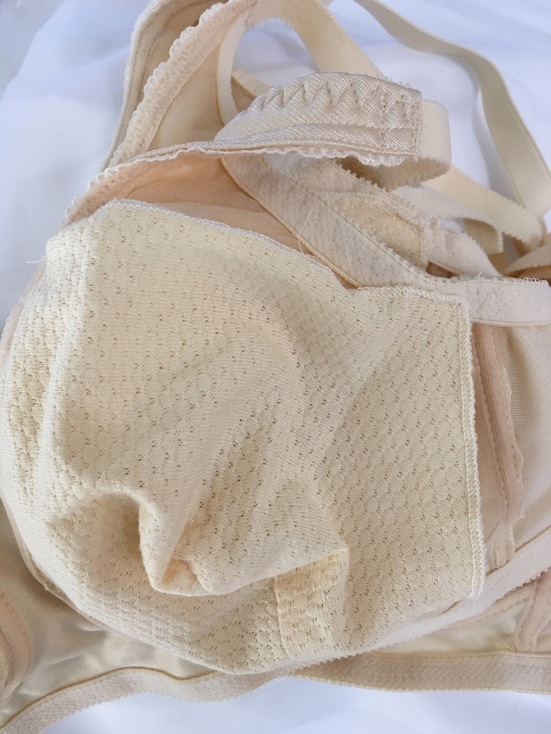 May include: A beige bra with adjustable straps and a textured, breathable cup design. The bra features a delicate lace trim along the edges and a soft, neutral colour, suitable for everyday wear. The image highlights the bra's construction and materials.