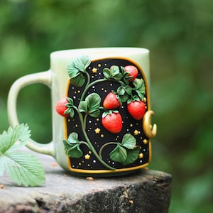 Può includere: Una tazza in ceramica verde chiaro con un motivo decorativo a fragole. Il design presenta fragole rosse, foglie verdi e uno sfondo nero con stelle dorate. Una luna crescente dorata è sul lato della tazza. Una foglia di fragola poggia sulla superficie di legno.