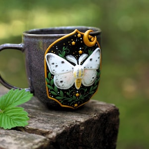 May include: A dark grey ceramic mug with a decorative plaque showing a white moth design. The plaque has a black background with gold stars and a crescent moon. Green leaves and a small strawberry plant are visible.
