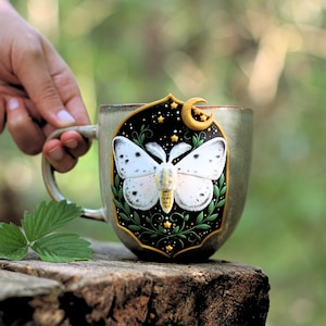 Może przedstawiać: Ceramiczny kubek z szczegółowym wzorem motyla. Motyl jest biały z czarnymi kropkami, na czarnym tle z gwiazdami i księżycem w kształcie półksiężyca. Kubek ma jasnozielony kolor i uchwyt.