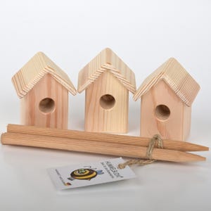 Könnte beinhalten: Drei kleine Vogelhäuschen aus Holz in einer Reihe, jedes mit einem runden Loch, durch das die Vögel eintreten können. Die Vogelhäuschen sind aus Naturholz gefertigt und haben ein schlichtes Design. Zwei Holzstiele sind zum Befestigen der Vogelhäuschen enthalten.