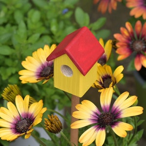 Könnte beinhalten: Ein kleines, gelbes Vogelhäuschen mit rotem Dach steht in einem Garten, umgeben von gelben Blumen mit violetten Zentren.