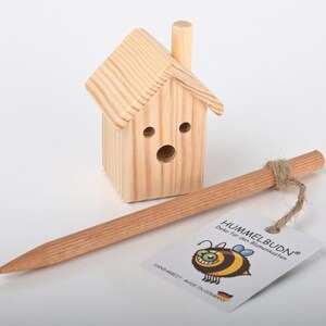 Könnte beinhalten: Ein kleines Holz-Bienenhaus mit Schornstein und einem runden Einflugloch. Es wird mit einem Holzstab zum Aufhängen geliefert. Das Etikett lautet "Hummelbudn® Deko für den Blumenkasten Handmade - Made in Germany".