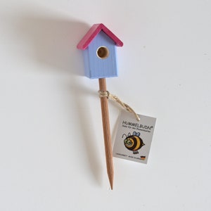 Könnte beinhalten: Ein kleines Vogelhaus aus Holz, hellblau mit rosa Dach, auf einem Stab montiert. Das Vogelhaus hat eine runde Öffnung und ist mit Schnur an einem Holzstab befestigt. Ein Etikett mit einer Cartoon-Biene und dem Text "HUMMELBUDN" ist angebracht.