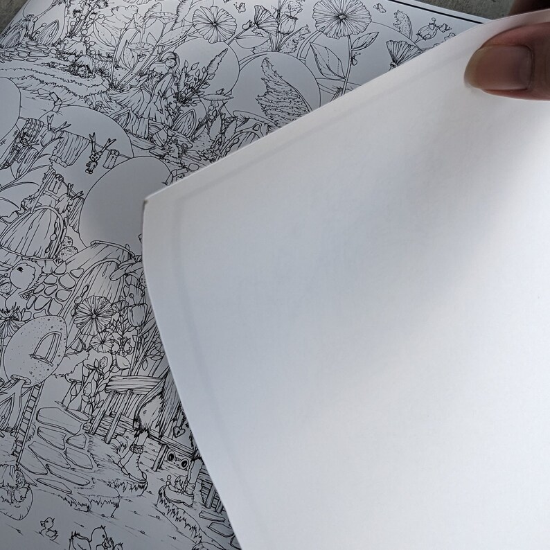May include: A partially visible coloring book page with a detailed black and white illustration of a whimsical garden scene. The page is being held open, revealing a blank white page. The artwork features flowers, houses, and other detailed elements.