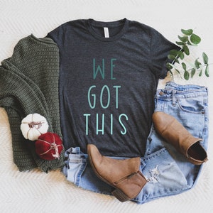 Puede incluir: Una camiseta gris oscuro con el texto "WE GOT THIS" en color verde turquesa. La camiseta est&aacute; colocada sobre una superficie blanca con un su&eacute;ter verde, jeans azules, botas marrones y peque&ntilde;as calabazas.