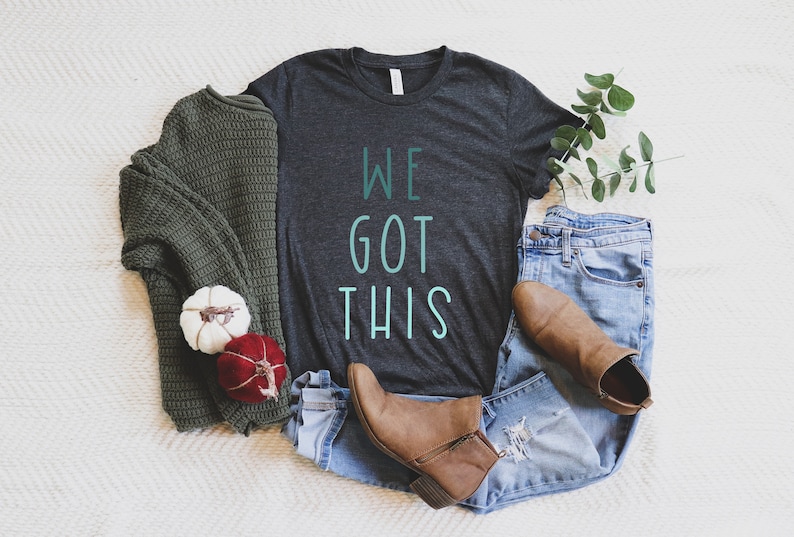 Puede incluir: Una camiseta gris oscuro con el texto "WE GOT THIS" en color verde turquesa. La camiseta est&aacute; colocada sobre una superficie blanca con un su&eacute;ter verde, jeans azules, botas marrones y peque&ntilde;as calabazas.