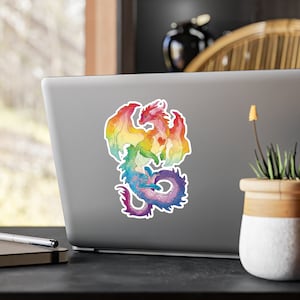 May include: A rainbow watercolour sticker of a dragon curled up with its wings spread out. The dragon is a vibrant mix of red, orange, yellow, green, blue, indigo, and violet.