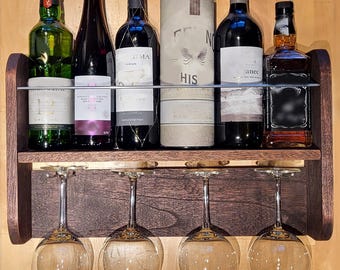 May include: A dark wood wine rack with four wine glasses suspended beneath. The rack holds six bottles of wine and spirits. The bottles vary in shape and size, with diverse labels. The rack is mounted on a light brown wooden wall.