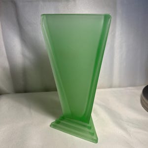 May include: A green glass vase with a triangular shape and a stepped base. The vase has a frosted finish and a wide opening. The design is reminiscent of Art Deco style. The vase is approximately 8 inches tall.