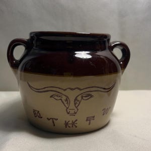 May include: A two-handled, brown and tan ceramic jar with a longhorn design. The jar has a dark brown rim and handles, with a lighter tan body. The longhorn design is etched in brown. The jar has a rustic, country aesthetic.