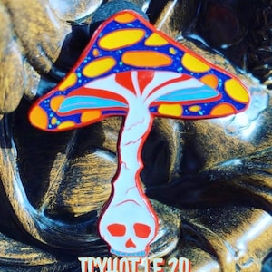 May include: A colorful enamel pin featuring a mushroom with a skull for a stem. The mushroom cap is red, blue, and yellow with white spots. The skull is white with red eyes. The pin has the text "ICYHOT LE 20" at the bottom.