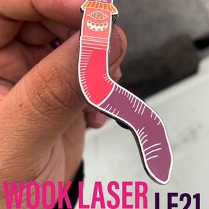 May include: A pink and purple enamel pin shaped like a worm with a hat and a single eye. The pin has a white outline and the text "Wook Laser LE21" is visible below the pin.