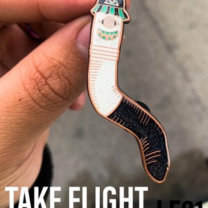 May include: A white and black enamel pin with a worm-like figure wearing a green and black hat. The pin has a sparkly finish and says "TAKE FLIGHT LE21" at the bottom.