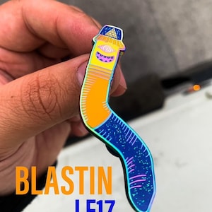 May include: A rainbow iridescent enamel pin shaped like a worm with a head that looks like a building with a triangle on top. The text "BLASTIN LE17" is visible in the image.