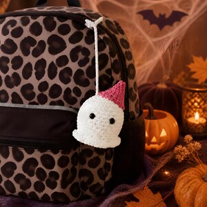 May include: A white crocheted ghost charm with a pink party hat and black bead eyes, attached by a white string to a gray and black leopard print backpack. The ghost features a rounded body and a wavy bottom edge, hanging from the zipper pull.