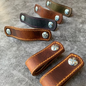 May include: Five leather straps with metal rivets and stitching. The straps are in different colors, including brown, black, and green. The straps are arranged in a row, with the brown straps at the front.