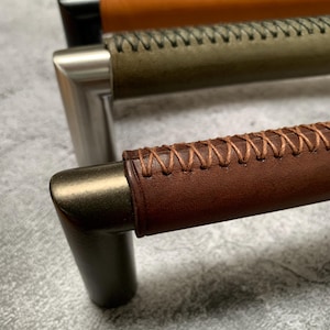 May include: Close-up of three cabinet pulls with metal hardware and leather-wrapped handles. The leather is stitched with a contrasting thread. Colours include brown, olive green, and orange.