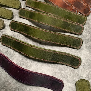 May include: Assortment of leather straps with contrasting stitching in various colours, including green, brown, purple, and blue. The straps are all hand-stitched and have a rustic, handmade look.