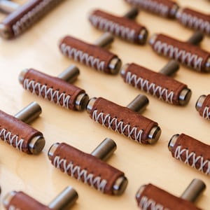 May include: A collection of brown leather cabinet knobs with white stitching. The knobs have a T-shaped design and are arranged in rows on a light wood surface.