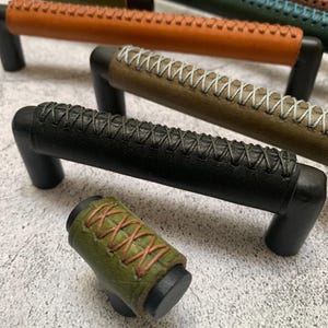 May include: A collection of cabinet pulls and a knob in various colors, including black, brown, green, and orange. The pulls have a leather-wrapped grip with decorative stitching. The hardware is made of black metal.