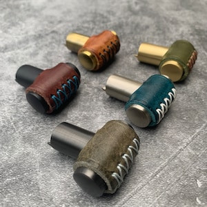 May include: Five different leather wrapped cabinet knobs with various colours of leather and stitching. The knobs are made of metal and have a T-shaped design.
