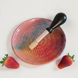 Könnte beinhalten: Eine rosa Keramikreibe mit einer Bürste und zwei frischen Erdbeeren. Die Platte hat eine strukturierte Oberfläche und farbige Akzente. Die Bürste hat einen schwarzen Griff und eine silberne Zwinge.