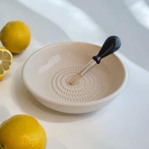 Könnte beinhalten: Eine cremefarbene Keramik-Reibeschüssel mit kreisförmigem Reibmuster und einem Pinsel mit schwarzem Griff. Zwei ganze Zitronen und eine Zitronenhälfte sind ebenfalls zu sehen, was auf ein kulinarisches Thema hindeutet.