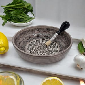 Könnte beinhalten: Graue Keramik-Reibeschüssel mit dekorativem Rand und Edelstahlreibe. Eine Bürste mit schwarzem Griff liegt in der Schüssel. Zitronen, Knoblauch und Grünzeug in der Nähe, was auf die Verwendung in der Küche hindeutet.