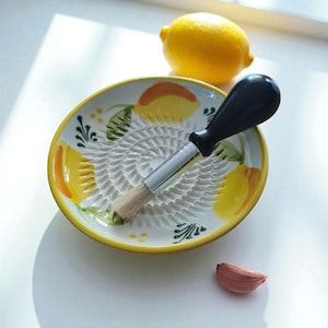 Könnte beinhalten: Eine Keramikreibe mit einem Pinsel mit schwarzem Griff, verziert mit gelben Zitronen und grünen Blättern. Eine ganze Zitrone und eine Knoblauchzehe sind ebenfalls vorhanden. Die Reibe hat eine weiße Oberfläche mit gelbem Rand.