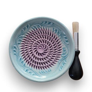 Könnte beinhalten: Ein hellblauer Keramik-Knoblauchschaber mit Spiralmuster und eine Bürste mit schwarzem Griff. Der Schaber hat einen dekorativen Rand und eine violette Mitte. Die Bürste hat einen Naturborstenkopf. Auf weißem Hintergrund.