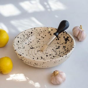 Könnte beinhalten: Eine weiße Keramikschüssel mit schwarzen Flecken und einem Knoblauchpressen-Design. Eine Knoblauchpresse mit schwarzem Griff sitzt in der Schüssel. Zwei ganze Knoblauchzehen und zwei Zitronen liegen auf einer weißen Oberfläche.
