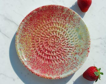 Könnte beinhalten: Ein Keramik-Reibeteller mit strukturierter Oberfläche und einem rot-gelb gesprenkelten Design. Zwei frische Erdbeeren sind in der Nähe des Tellers platziert. Der Teller hat eine zentrale Reibefläche und einen hellgrünen Rand.