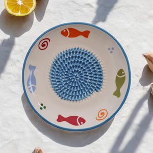 Könnte beinhalten: Ein weißer Keramik-Knoblauchpresseteller mit einer blauen, strukturierten Mitte und bunten Fischdesigns am Rand.