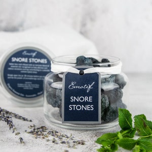 May include: A glass jar filled with dark blue and green stones labelled "Emotif Snore Stones". The jar is sitting on a white surface with a white towel and lavender flowers in the background.