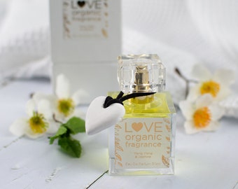 May include: A clear glass bottle of Love organic fragrance with a white heart charm. The bottle is filled with a yellow liquid and has a gold cap. The label reads "Love organic fragrance, Ylang Ylang & Jasmine, Eau de Parfum 50 ml."