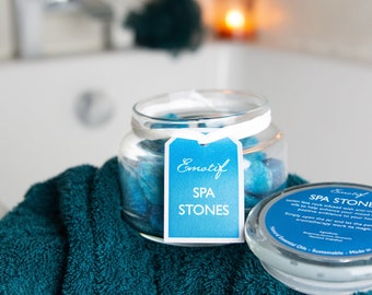 May include: A glass jar filled with blue spa stones, labeled "Emotif SPA STONES". The jar is sitting on a teal towel. The label on the jar reads "Emotif SPA STONES". The jar is also labeled "SPA STONE Italian lava rock infused with anti-vie oils to help enhance your mood and positive ambience to your home. Simply open the jar and let the powerful aromatherapy work its magic. Natural Essential Oils Sustainable Made in"