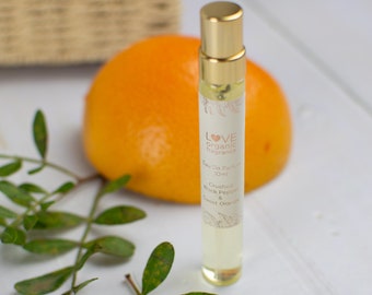 May include: A small, clear glass bottle with a gold cap and a label that reads "LOVE organic fragrance Eau De Parfum 10ml Crushed Black Pepper & Sweet Orange". The bottle is sitting on a white surface with a green leaf and a bright orange.