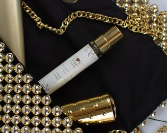 May include: A small, clear glass bottle of Love Organic fragrance in Vanilla & Topa. The bottle is in a black purse with a gold chain and gold embellishments.