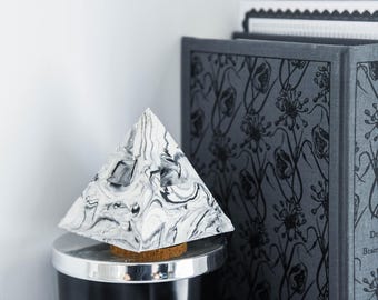 May include: A white and black marbled pyramid-shaped object sits on top of a black cylindrical container. The container is in front of a book with a black and white floral pattern and the text "Dracula Bram Stoker".