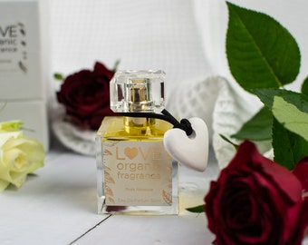 May include: A clear glass bottle of Love Organic fragrance with a gold label and a white heart charm. The bottle is sitting on a white wooden surface with red roses and white flowers.