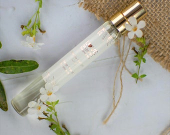 May include: A small, clear glass bottle of Love & Gardenia fragrance oil with a gold cap. The bottle is sitting on a white surface with white flowers and green leaves.