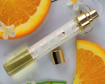 May include: A small glass bottle of orange blossom scented perfume with a gold cap. The bottle is labeled "Love Organic Fragrances" and "Eau de Parfum" with a floral design. The bottle is sitting on a white surface with orange slices and white flowers.