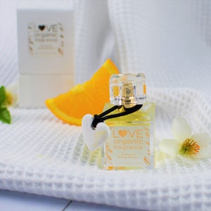 May include: A clear glass bottle of Love Organic fragrance with a black ribbon and a white heart charm. The bottle is labelled "Love Organic fragrance" and "Petal & Orange Blossom". The bottle is sitting on a white towel with a slice of orange and a white flower.
