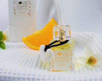 May include: A clear glass bottle of Love Organic fragrance with a black ribbon and a white heart charm. The bottle is labeled "Love Organic fragrance" and "Petal & Orange Blossom". The bottle is sitting on a white towel with a slice of orange and a white flower.