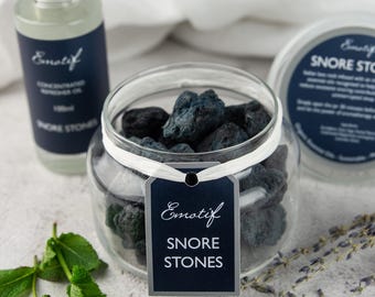 May include: A glass jar filled with dark gray lava rocks, labeled "Emotif Snore Stones". The jar is sitting on a white surface with a small sprig of green mint leaves and lavender flowers.