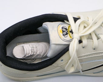 May include: A white leather sneaker with black accents and a yellow and black logo that says "Tennis Classic" inside the shoe. The laces are white and tied in a bow.