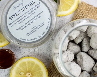 May include: A glass jar filled with gray lava rocks, labeled "Stress Stones" with a list of essential oils including tea tree, eucalyptus, petitgrain, sage, manuka, honey, lemon, hyssop, peppermint, and ginger. The jar is surrounded by lemons and a brown burlap surface.