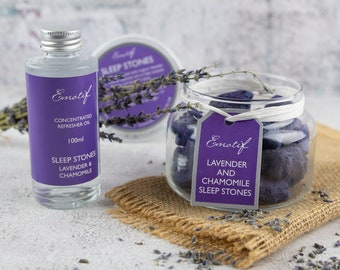 May include: A set of lavender and chamomile sleep stones in a glass jar with a white ribbon and a purple tag that reads "Emotif Lavender and Chamomile Sleep Stones". A clear glass bottle with a silver lid and a purple label that reads "Emotif Concentrated Refresher Oil 100ml Sleep Stones Lavender & Chamomile" is next to the jar.  Dried lavender flowers are scattered around the jar and bottle.